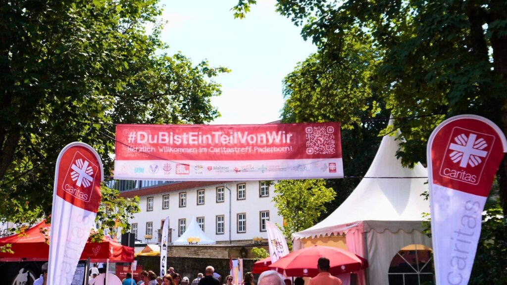 Das Bild zeigt einen festlichen Außenbereich mit mehreren Caritas-Logos und einem großen Banner, auf dem steht: „#DuBistEinTeilVonWir – Herzlich Willkommen im Caritastreff Paderborn“. Im Hintergrund sind Bäume, ein Gebäude und mehrere Zelte zu sehen, die Teil einer Veranstaltung zu sein scheinen. Der Bereich wirkt einladend und gut besucht, und der Slogan hebt die Gemeinschaftsarbeit der Caritas hervor.