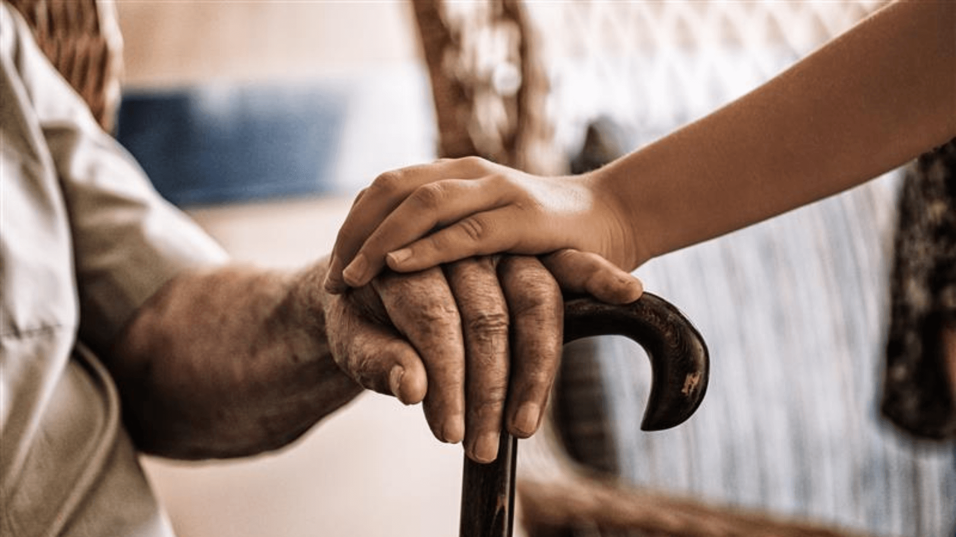 Eine jüngere Hand liegt tröstend auf der älteren, von der Arbeit gezeichneten Hand eines Menschen, die sich auf einen dunklen Gehstock stützt. Die Aufnahme ist in warmen, sepia-ähnlichen Tönen gehalten und vermittelt einen Moment der Fürsorge und des Beistands.