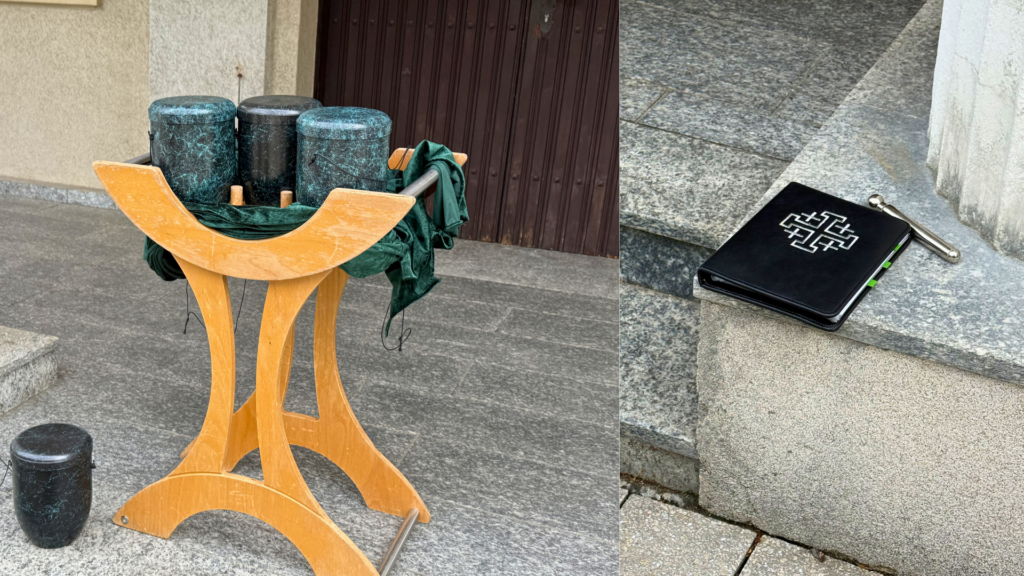 Holzgestell vor einem Gebäude mit drei dunklen Urnen, grünem Tuch und weiterem Gefäß am Boden, daneben ein Buch mit Kreuzsymbol und ein Stab auf einer Steinstufe.
