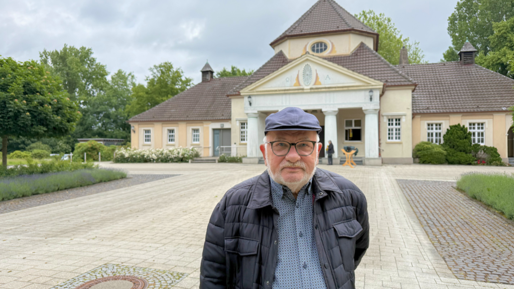 Person steht auf einem gepflasterten Vorplatz vor einem Gebäude mit Säulen und Eingangsbereich, im Hintergrund Bäume und Wege.